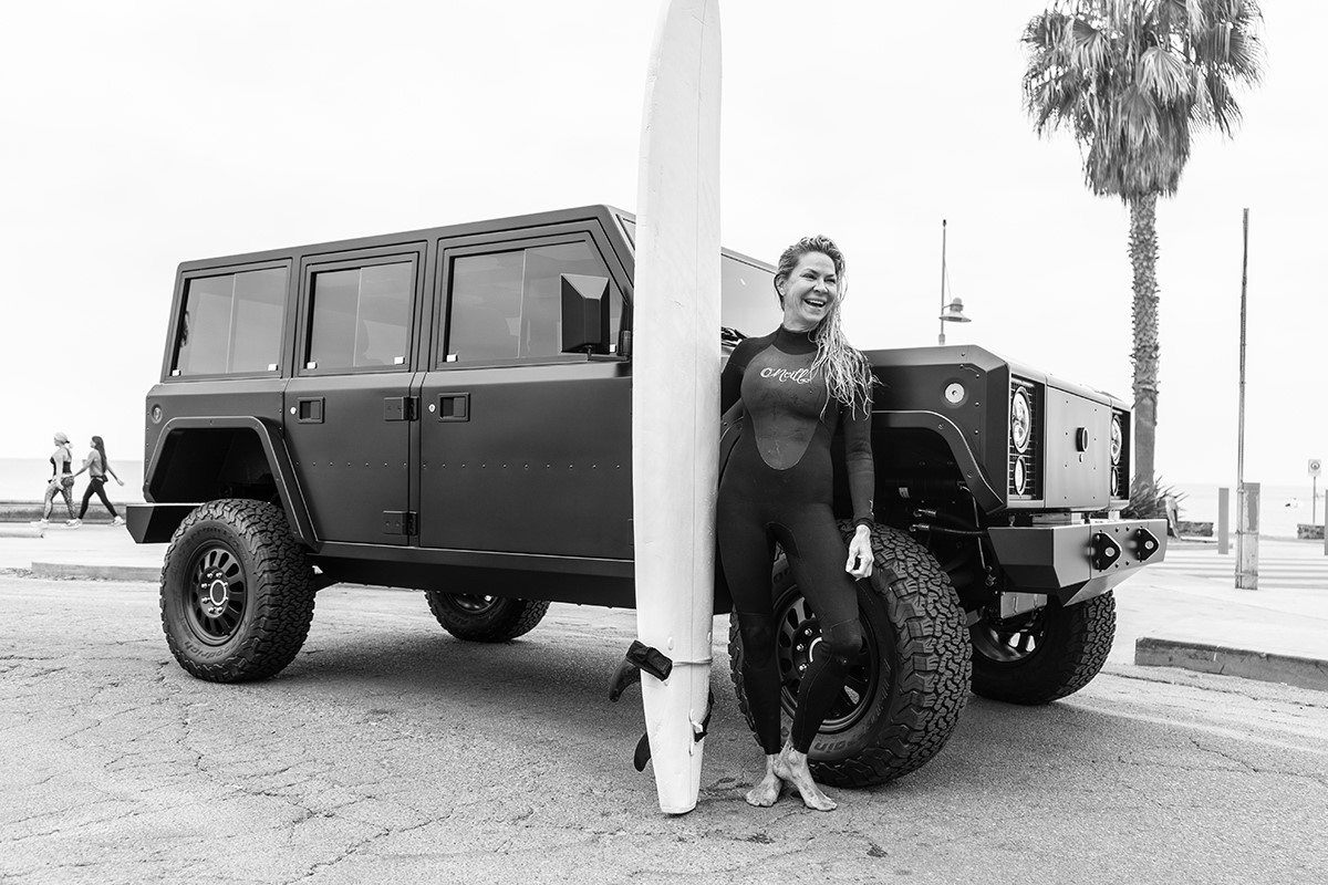 A woman surfer with board standing by the Bollinger B1 in California.